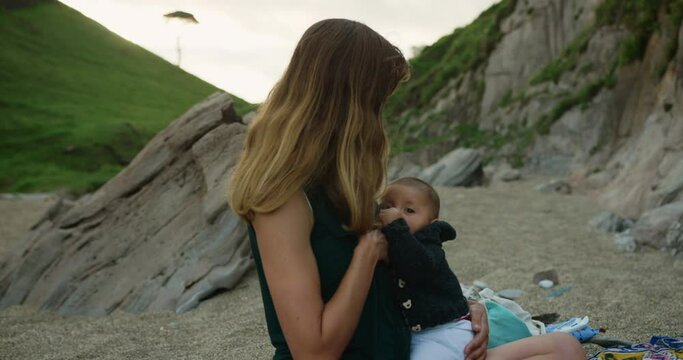 A young mother is breastfeeding her baby on the beach in summer at sunset