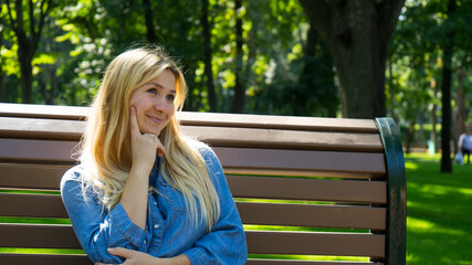 Portrait of attractive casual blond girl thoughtfully looking away and thinking on something  sitting in the sunny city park. Pretty young thoughtful woman confused about something sits on bench