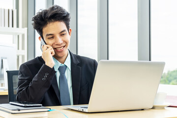 Smiling young asian businessman sitting at desk  talking on mobile phone and using laptop in workplace