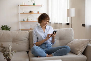 Obraz premium Young Caucasian woman sit on comfortable couch in living room text message on modern cellphone gadget. Female relax on sofa at home browse wireless internet on device or shop online.