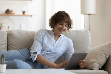 Smiling young Caucasian woman sit rest on couch in living room talk on video call on tablet gadget. Happy female relax on sofa at home browse use internet, have webcam virtual conversation.