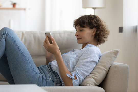 Young Caucasian woman relax on sofa in living room browsing wireless internet on smartphone. Female rest on couch at home texting messaging on modern cellphone gadget, use new technologies.