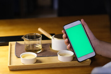over shoulder view of people holding green screen smart phone with traditional Chinese tea set