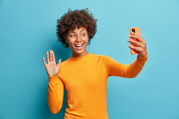 Pretty curly haired positive woman holds modern mobile phone and makes video call waves palm in hello gesture wears casual jumper isolated over blue background. People modern technology concept © wayhome.studio