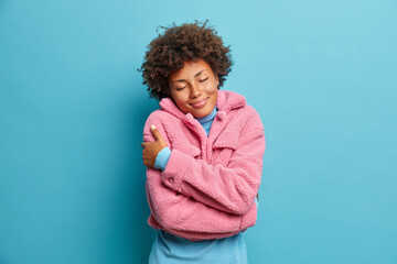 Photo of pleased beautiful Afro American woman enjoys comfort in new warm rosy coat embraces herself and stands with closed eyes recalls pleasant memories feels pleased during cold winter day - obrazy, fototapety, plakaty