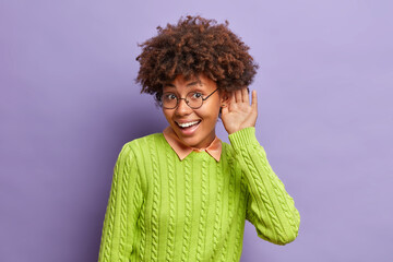 Happy curious dark skinned woman holds and near ear and to overhear interesting informaion wears casual green jumper stands against purple background. I want to hear everything. Lady eavesdropes