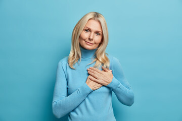 Beautiful blonde adult woman makes gratitude gesture presses hands to heart being thankful for compliment appreciates something wears casual turtleneck poses over blue background. Body language - obrazy, fototapety, plakaty