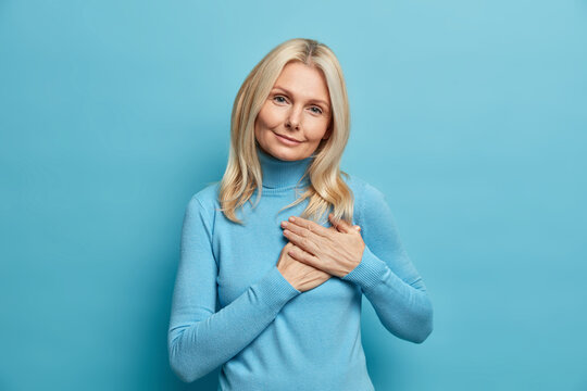 Beautiful blonde adult woman makes gratitude gesture presses hands to heart being thankful for compliment appreciates something wears casual turtleneck poses over blue background. Body language