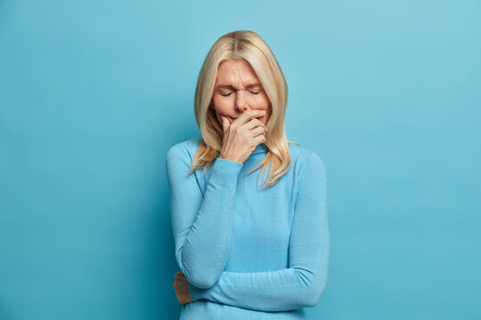 Troublesome blonde middle aged European woman whins and complains about something feels unwell worries about her children dressed in casual blue turtleneck poses indoor. Life problems concept