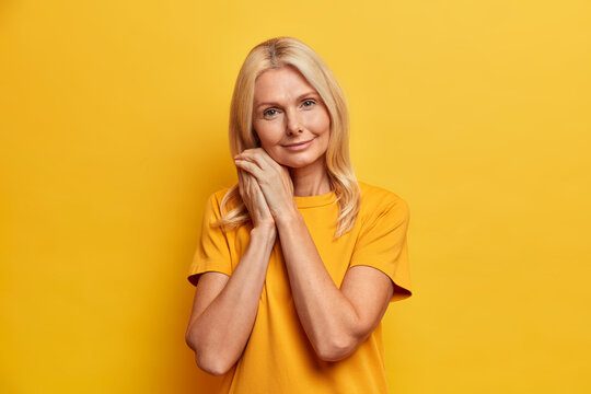 Portrait of good looking senior woman looks confidently at camera leans at hands dressed in casual yellow t shirt poses indoor. Delighted gorgeous fifty years old lady stands relaxed indoor.