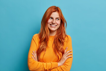 Charming positive redhead European woman stands crossed hands indoor, smiles gently with teeth and...