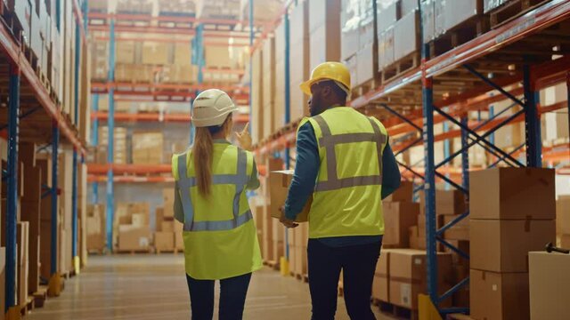 Male and Female Workers Wearing Hard Hats Walking Through Retail Warehouse full of Shelves with Goods in Cardboard Boxes. Working in Logistics and Delivery Center. Following Slow Motion Shot