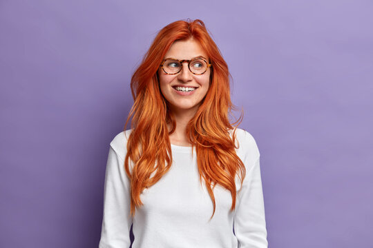 Portrait of smiling good looking young woman has ginger hair, looks aside and wears casual white jumper. Charming young millennial girl looks positively, stands against vivid purple background