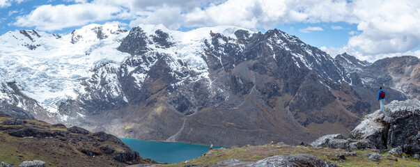 Panorama landscape of man standing in a rock during hike/trail with huge mountains with snow and a lake with blue sky and clouds, in cordilera Huaytapallana, Huancayo, peru