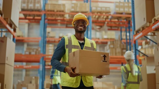 Handsome Male Worker Wearing Hard Hat Holding Cardboard Box Walking Through Retail Warehouse full of Shelves with Goods. Working in Logistics and Distribution Center. Front Following Slow Motion 