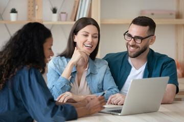 African American manager realtor consulting happy couple at meeting, using laptop, making presentation, sitting at table in modern office, smiling wife and husband taking loan or mortgage