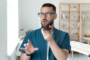 Close up confident young businessman wearing glasses recording voice message, using smartphone in office, serious employee chatting online or speakerphone, activating digital assistant