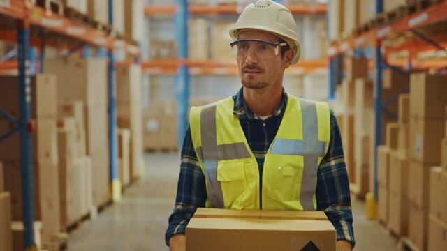Handsome Male Worker Wearing Hard Hat Holding Cardboard Box Walking Through Retail Warehouse full of Shelves with Goods. Working in Logistics and Distribution Center. Front Following Slow Motion Shot
