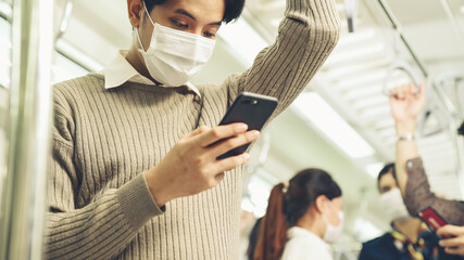 Traveler wearing face mask while using mobile phone on public train . Coronavirus disease or COVID 19 pandemic outbreak and urban city lifestyle problem in rush hour commuting concept .