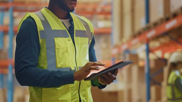 Male Worker Wearing Hard Hat Checks Products Stock and Inventory with Digital Tablet Standing in Retail Warehouse full of Shelves with Goods. Distribution, Logistics. Zoom in on Tablet Computer