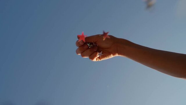 Woman hand drops silver stars sequins, confetti on top of the camera. relaxing, naive, romantic, creative Slow motion video footage. Blue summer sky on the background. party dreams time concept mood