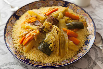 Top view food shot of a typical moroccan couscous served on a family size ceramics tagine plate surrounded by glasses, sauce, spoons and spices on a fine linens background. Blessed friday concept.