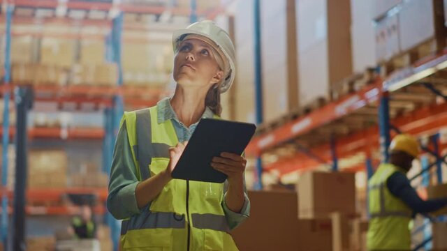 Professional Female Worker Wearing Hard Hat Checks Stock and Inventory with Digital Tablet Computer in the Retail Warehouse full of Shelves with Goods. People Working in Logistics Distribution Center 