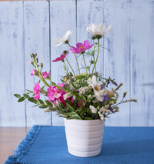 multicolored wildflowers stand on a wooden table, on a blue cloth napkin, in a white vase against a blue wall    