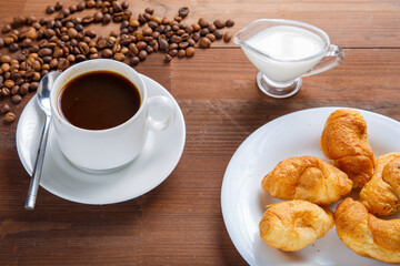 A cup of coffee on the table next to sugar and cream in a jug and a croissant.