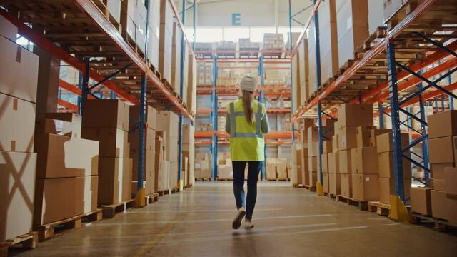 Professional Female Worker Wearing Hard Hat Checks Stock and Inventory Walks in the Retail Warehouse full of Shelves with Goods. Working in Logistics, Delivery Center. Following Back View 