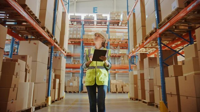 Professional Female Worker Wearing Hard Hat Uses Digital Tablet Checks Inventory Walks in the Retail Warehouse full of Shelves with Goods. Working in Logistics, Distribution Center. Following Shot 