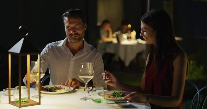 Happy elegantly dressed couple is enjoying romantic dinner together and cheering with white wine glasses to celebrate their anniversary and timeless love at well-served table at the luxury restaurant