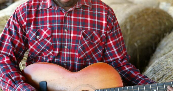 Close up of African American young handsome man sitting on hay stocks in shed and playing music on guitar. Male farmer singing and play on musical instrument song. Countryside concept. Village.