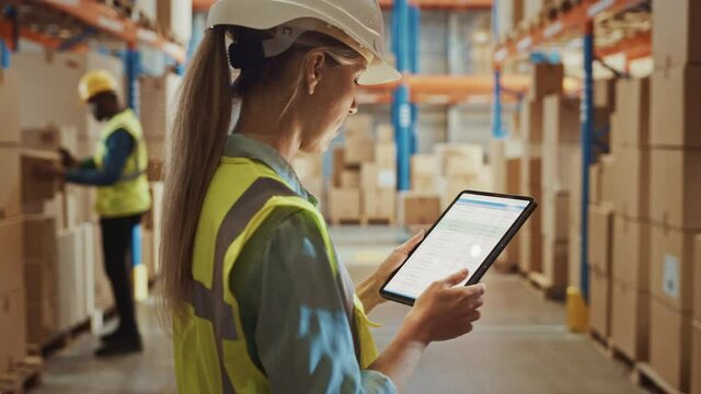 Professional Female Worker Wearing Hard Hat Uses Digital Tablet Computer with Inventory Checking Software in the Retail Warehouse full of Shelves with Goods. Delivery, Distribution Center. Side View 