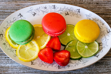 Looking down at a plate of French macron cookies, red, yellow and green with fresh lemon and lime slices and fresh cut strawberries