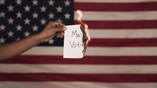Hands holding burning Mail-in voting paper with US flag as background - Concept against the mail by vote in US Election