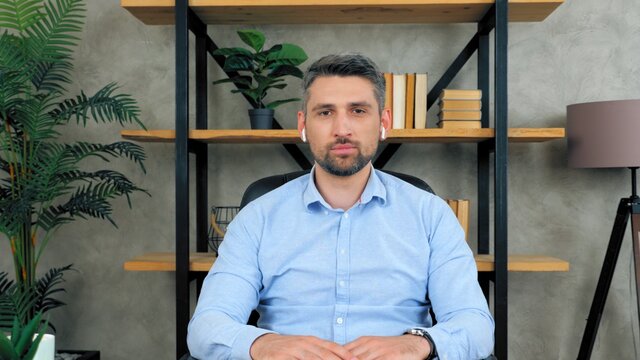 Confident business man in home office wearing wireless earphones looks camera listens conducts remote distant online interview potential employee to company by video chat conference, web cam concept