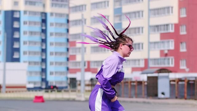 Young beautiful woman with colored pigtails dancing hip hop outdoors on the background of high-rise buildings, jumping cheerfully and looking at the camera. Slow motion