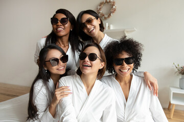 Selfie camera view shot beautiful young multiracial diverse girl besties in white silk gowns wearing dark sunglasses, having fun together at bridal bachelorette birthday spa party together indoors.
