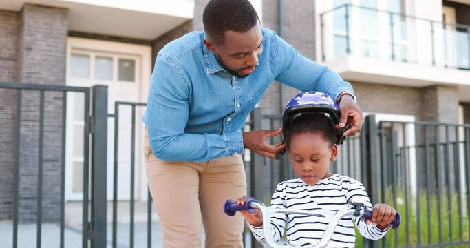 African American father wearing helmet on small pretty girl and teaching ride on bike at street. Little kid in casque learning riding bicycle. Schoolgirl and daddy. Daughter sitting on vehicle.