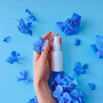 Top View And Close-up Of White Plastic Spray Bottle Mockup, Female Hand And Blue Flowers On A Blue Background. Natural Organic Spa Cosmetics And Liquid Antimicrobial Spray Concept. Square Flatlay.