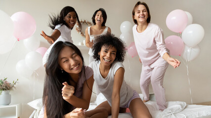Overjoyed attractive young diverse multiracial girls in sleepwear enjoying funny birthday photoshoot at home sleepover party, posing on bed, laughing joking having fun entertaining together indoors.