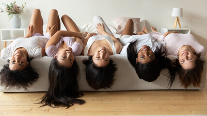 Happy playful multiracial diverse brunette girls besties lying on bed upside down, having fun...