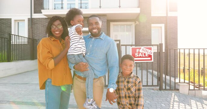 Portrait of happy African American family with small children standing at new house at suburb and demonstrating keys to camera. Cheerful parents and kids showing key moving in new home. Sold.