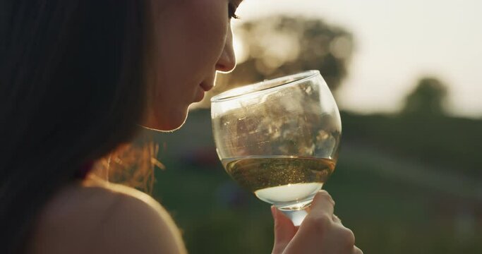 Authentic shot of happy young elegantly dressed woman is tasting a flavor and drinking a white wine poured in transparent glass on scenic vineyards background at sunset.