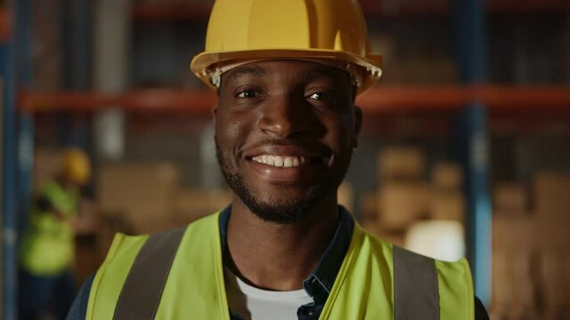 Handsome and Happy Professional Worker Wearing Safety Vest and Hard Hat Charmingly Smiling on Camera. In the Background Big Warehouse with Shelves full of Delivery Goods. Medium Close-up Portrait