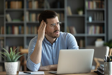 Unhappy young Caucasian male worker sit at desk at home office distracted from computer work look...