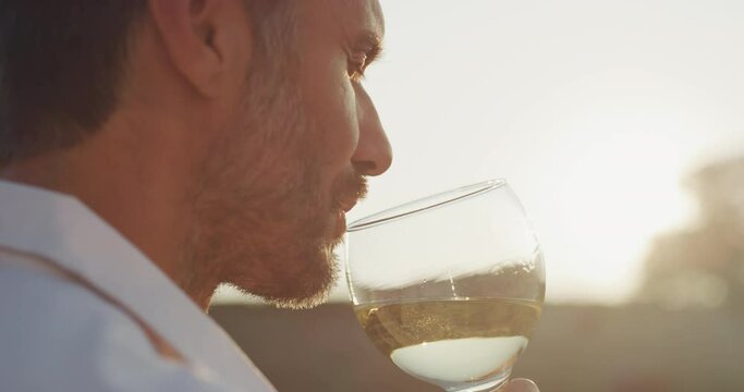 Authentic shot of happy handsome elegantly dressed man is tasting a flavor and drinking fresh white wine poured in transparent glass on scenic vineyards background at sunset.