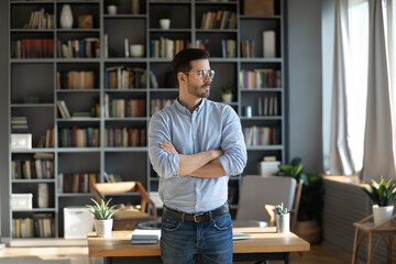 Obraz premium Serious confident young 30s Caucasian businessman stand near home office desk look in distance thinking or planning. Pensive millennial male pondering making decisions. Business vision concept.