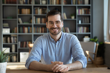 Headshot portrait of smiling 30s Caucasian millennial man in glasses sit at desk at home talk speak on video call or webcam virtual conference. Picture of happy young male in spectacles pose at table. - obrazy, fototapety, plakaty